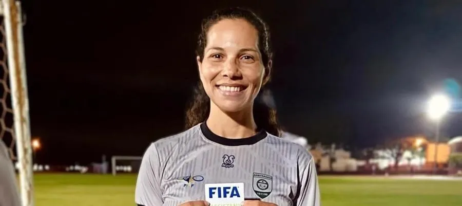 A árbitra Leila Cruz, de uniforme da FIFA, sorri em campo de futebol à noite