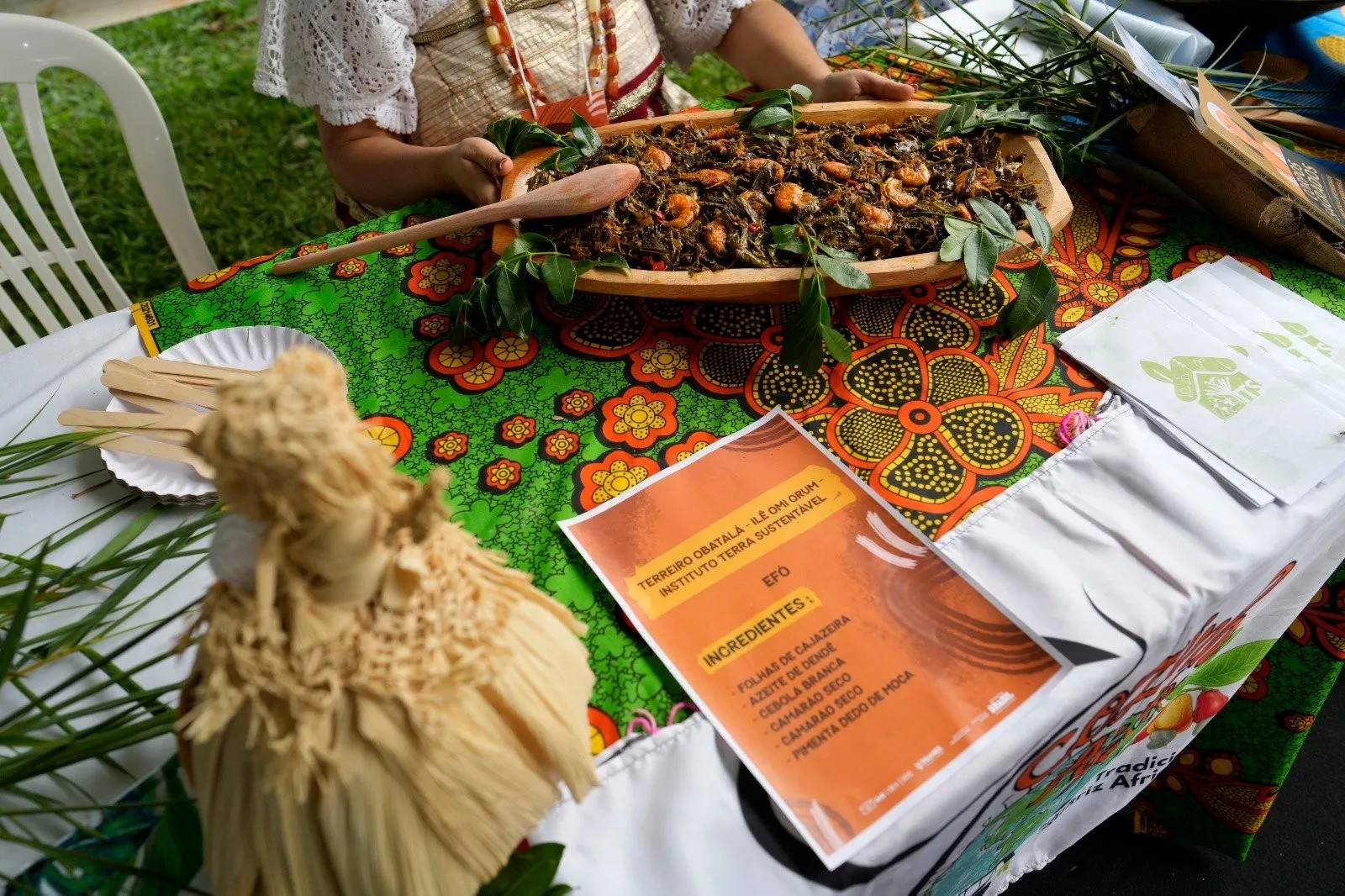 Travessa de madeira com efó sobre mesa forrada com tecido estampado, ao lado de ficha com os ingredientes do prato