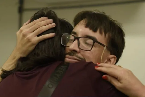 Homem de óculos abraça uma pessoa, de olhos fechados