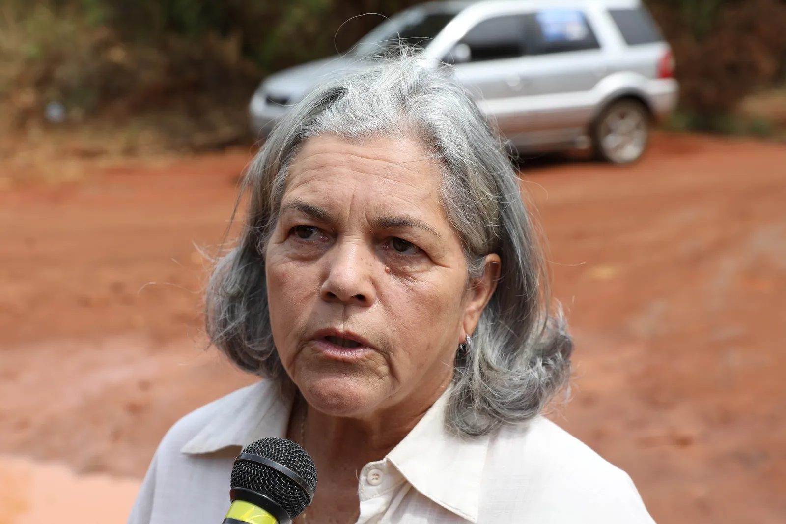 Tereza Cristina Souza Pires, presidente de associação de moradores, fala ao microfone diante de uma estrada de terra