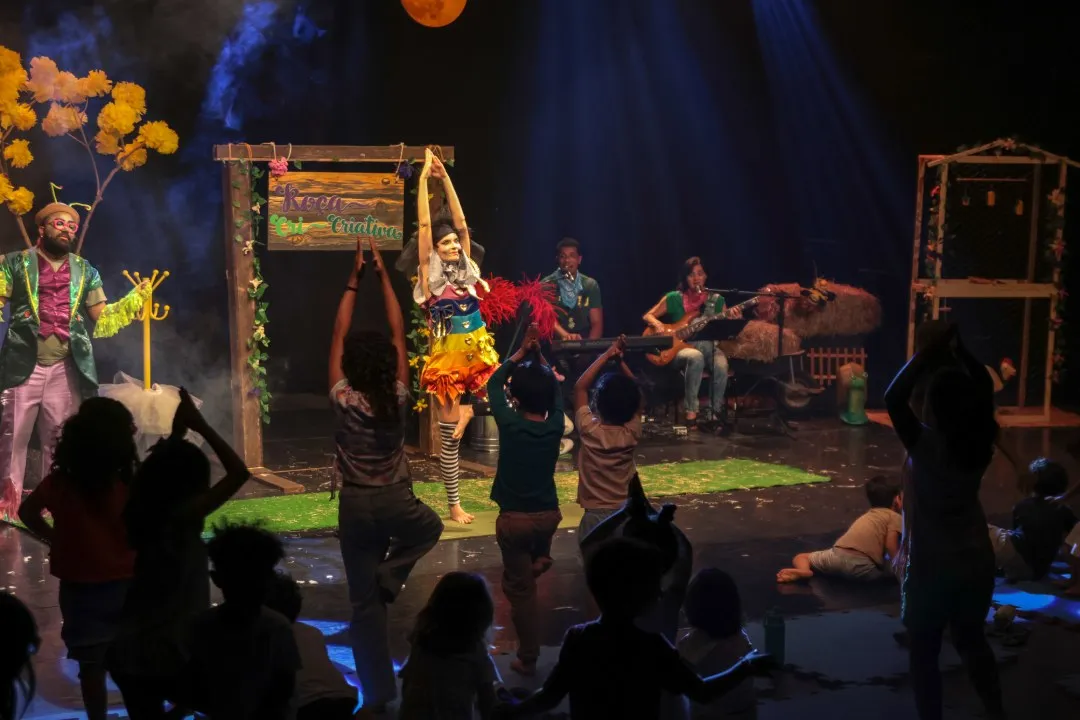 Cena do musical Roça Cri-criativa: atores fantasiados no palco iluminado, com crianças da plateia de costas em primeiro plano