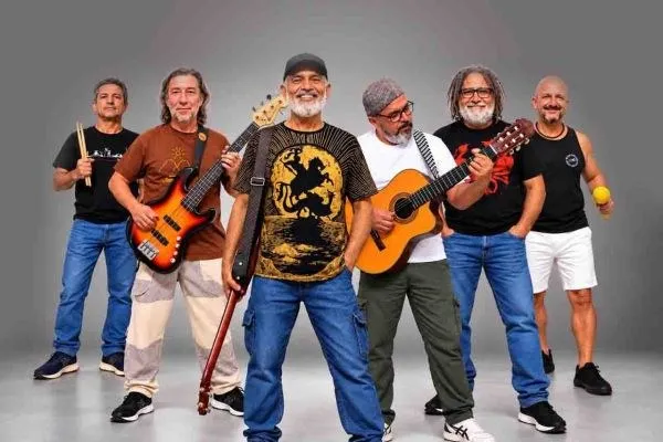 Os seis integrantes da Banda Pra Quem Tá Quente posam de pé com guitarra, violão e baquetas diante de um fundo cinza