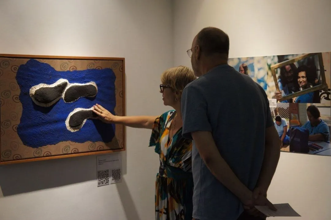 Duas pessoas tocam um quadro tátil azul da exposição Sob os Pés do Mundo